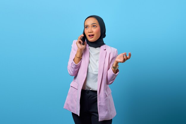 Surprised Asian woman talking with friend by mobile phone over blue background