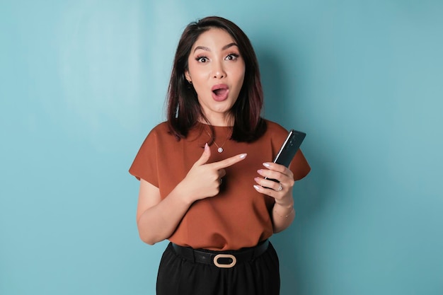 Удивленная азиатка в коричневой рубашке держит свой смартфон на синем фоне