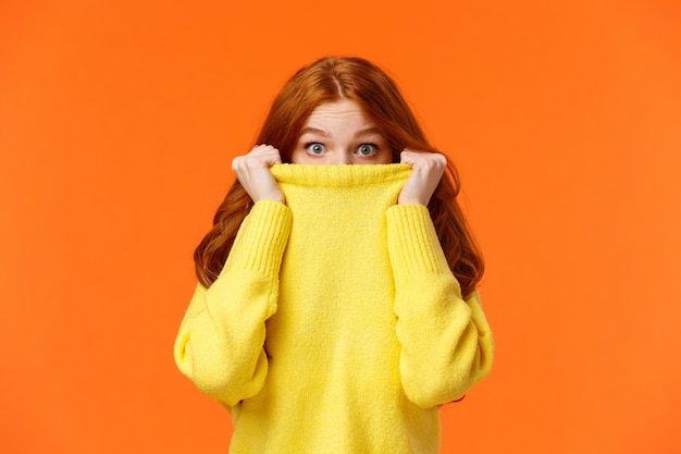 Surprised and astonished cute curious redhead female pulling sweater collar on face and popping eyes