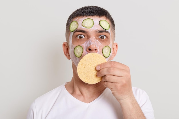 Surprised astonished man wearing white t shirt doing cosmetic procedures at home with mask slices of cucumber looking at camera and covering mouth with sponge posing isolated over gray background