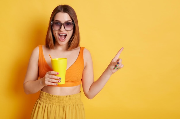 Donna stupita sorpresa che indossa una parte superiore arancione in piedi isolata su sfondo giallo che tiene la tazza con il bere che punta lontano nell'area pubblicitaria