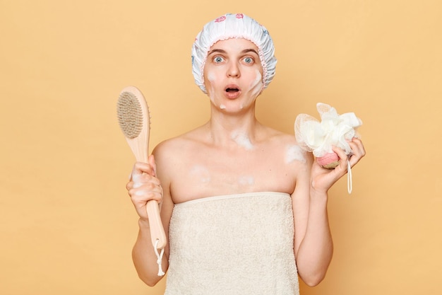 Surprised astonished woman wearing shower cap standing isolated over beige background holding washcloth and brush for dry massage