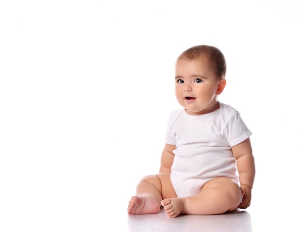 Surprised baby kid sitting on white studio floor. Inquisitive child wearing jumpsuit with confused expression on cute face looking at camera. Isolated caucasian toddler portrait