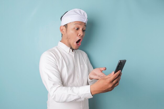 Удивленный балийский мужчина в уденге или традиционной повязке на голове и белой рубашке держит свой смартфон на синем фоне