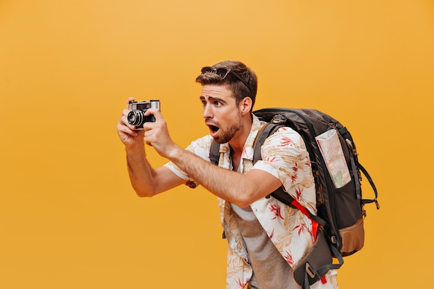 Surprised bearded cool man with stylish hairstyle in printed white summer shirt and grey tshirt taking photo and posing with large backpack