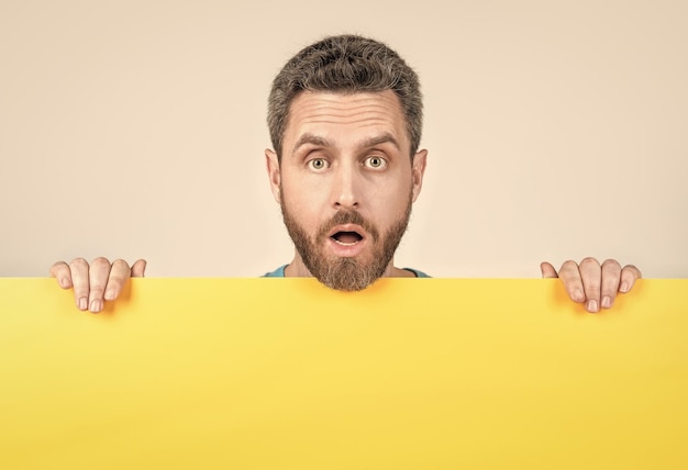 Surprised bearded man behind blank yellow paper banner with\
copy space for marketing design promo