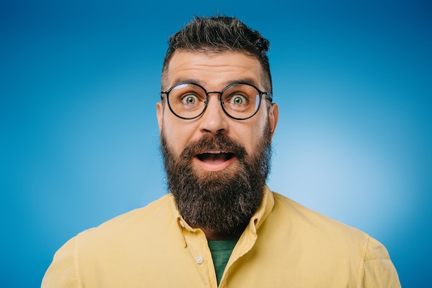 Surprised bearded man in eyeglasses looking at camera isolated on blue