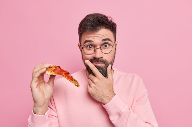 Surprised bearded man holds slice of pizza eats fast food has tasty snack for good mood wears round spectacles casual long sleeved jumper 