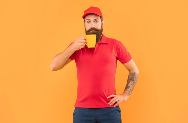 写真 マグカップの黄色の背景のコーヒーを保持しているカジュアルな赤い帽子とtシャツで驚いたひげを生やした男