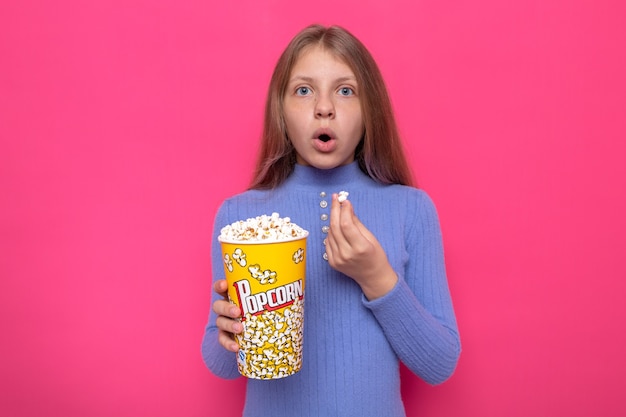 Photo surprised beautiful little girl wearing blue sweater holding popcorn bucket