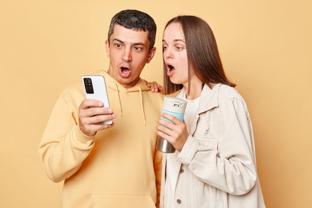 Surprised beautiful woman and handsome man wearing casual style clothing standing isolated over beige background browsing internet together on smartphone drinking hot coffee from thermos
