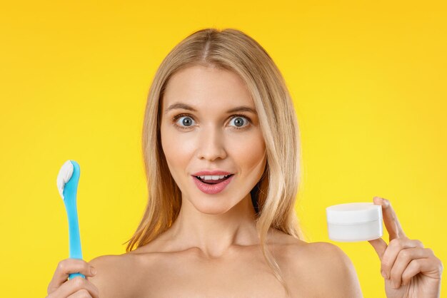 Surprised beautiful woman holding skin cream over yellow background