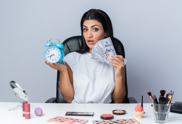 Surprised beautiful woman sits at table with makeup tools holding alarm clock with cash 