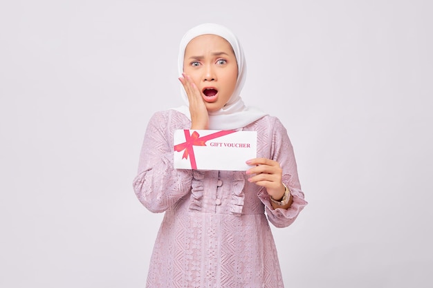 Photo surprised beautiful young asian muslim woman wearing hijab and purple dress holding gift voucher certificate and looking at camera isolated on white studio background