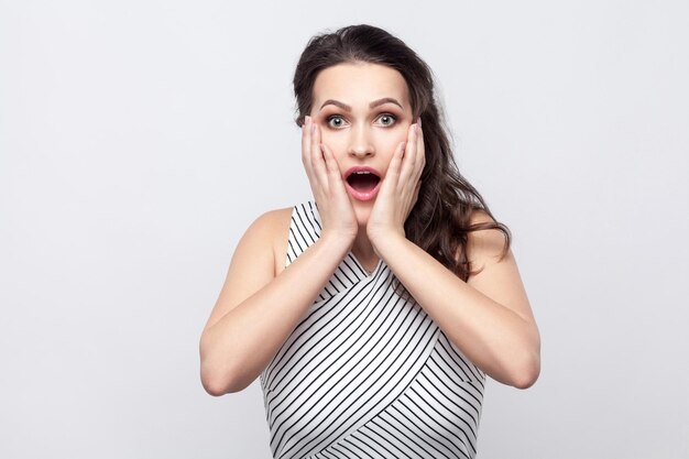 Surprised beautiful young brunette woman with makeup and striped dress standing touching her face and looking at camera with unbelievable face. indoor studio shot, isolated on grey background.