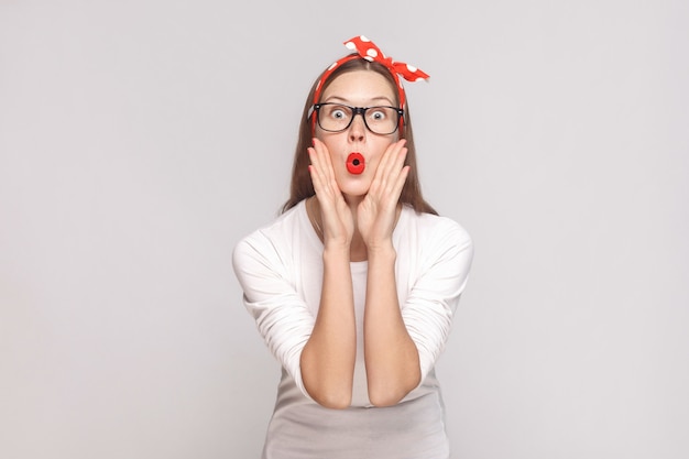 Surprised big eyes portrait of beautiful emotional young woman in white t-shirt with freckles, black glasses, red lips and head band. indoor studio shot, isolated on light gray background.