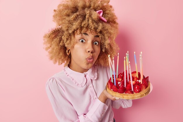 Surprised birthday girl looks with wondered expression holds\
delicious strawberry cake with burning candles makes wish\
celebrates anniversary dressed in blouse isolated over pink\
background