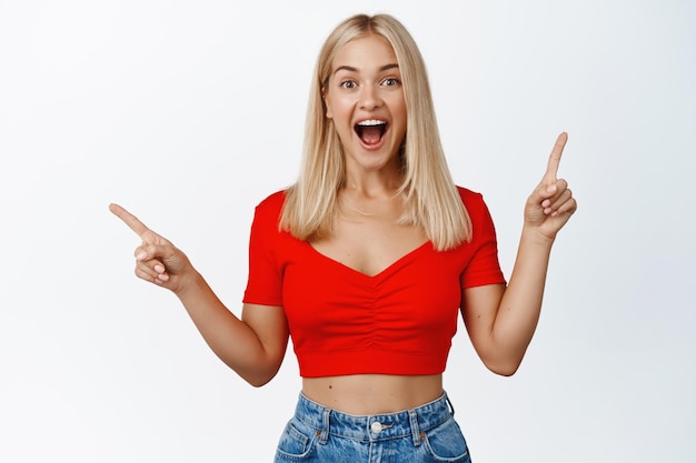 Surprised blond girl shows two options points sideways and smiling amazed excited with news stands against white background