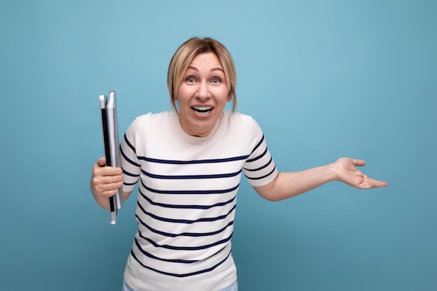 Surprised blond girl student in a casual outfit with a laptop in her hands on a blue background