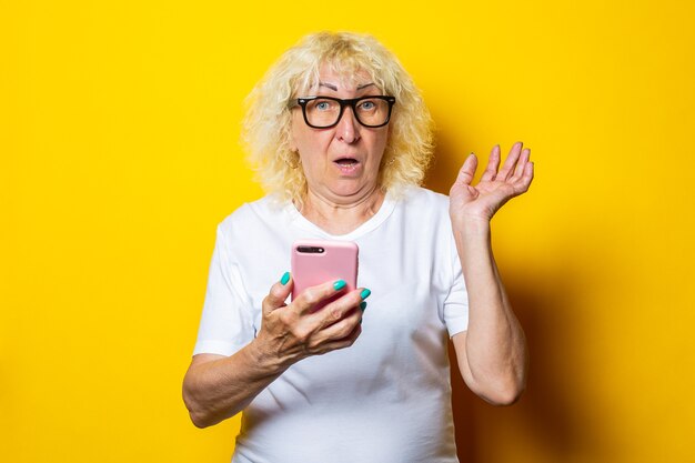 Surprised blonde old woman in white t-shirt and glasses holding phone