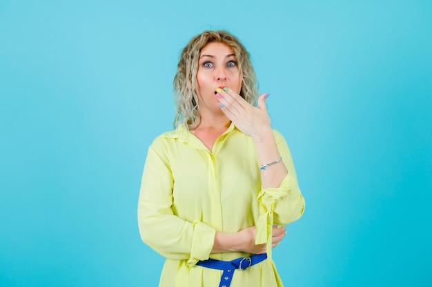 Surprised blonde woman is looking at camera by holding hand on mouth on blue background