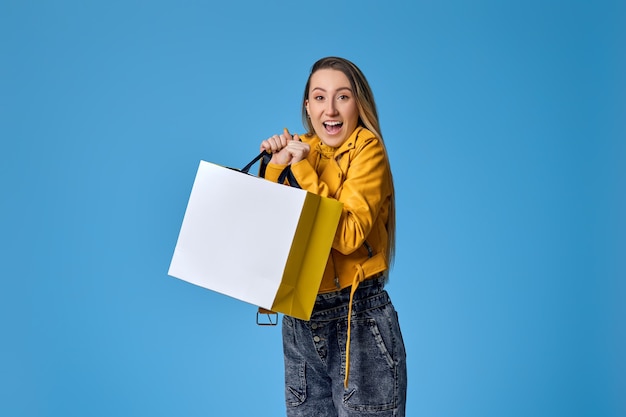 Surprised blonde woman in leather jacket holding shopping bag with purchases on blue background. Mock up.