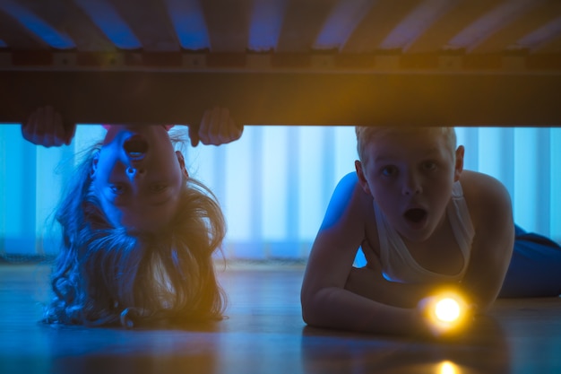 The surprised boy and girl with a flashlight look under bed. night time