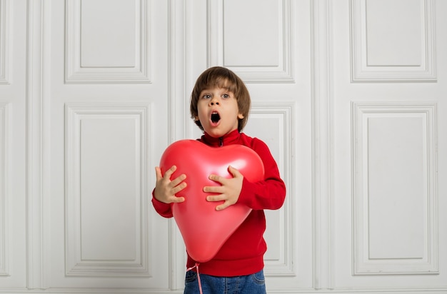 A surprised boy in jeans and a sweater holds a red heart balloon on a white background with space for text