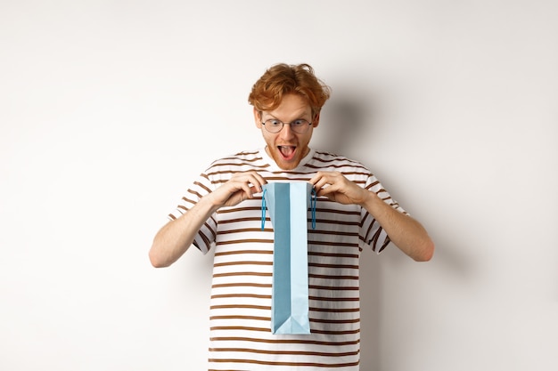 놀란 남자친구는 발렌타인 데이 선물로 쇼핑백을 열고 놀란 얼굴로 안을 쳐다보고 흰색 배경 위에 서 있습니다.
