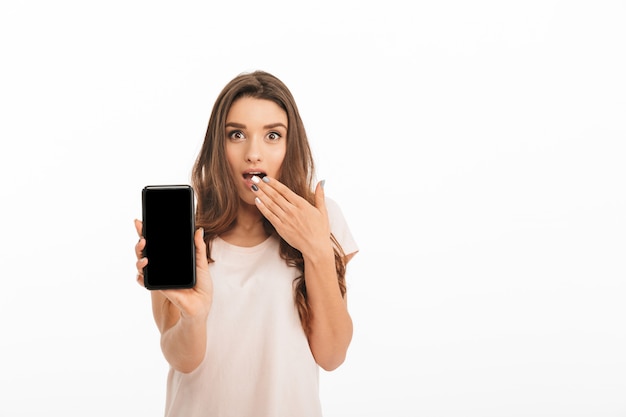 Surprised brunette woman in t-shirt showing blank smartphone screen over white wall