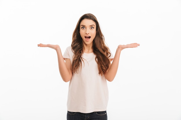 Surprised brunette woman in t-shirt shrugs her shoulder  over white wall