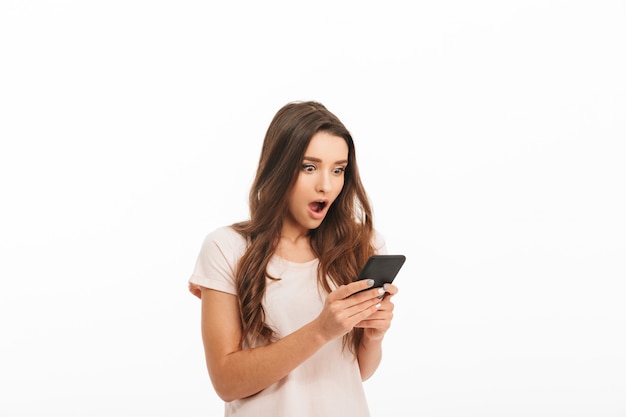 Surprised brunette woman in t-shirt writing message on smartphone over white wall