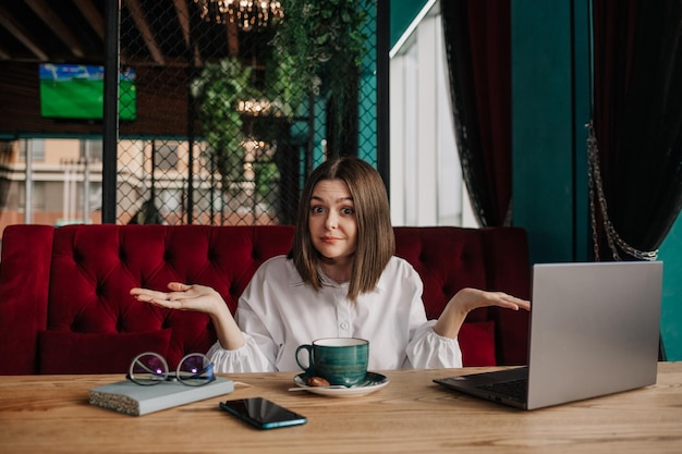 A surprised business woman is sitting at a table in a cafe with a cup of coffee and working on a laptop