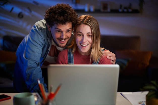 驚いた白人のカップルが暗いリビングの机に座ってコンピューターを見て幸せに見えます
