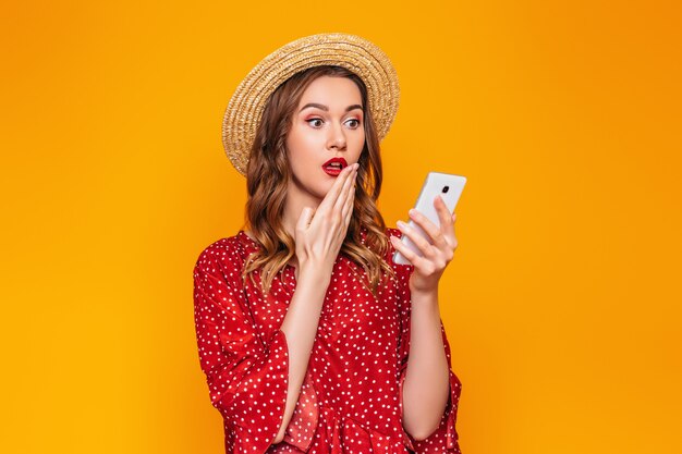 A surprised caucasian girl covers her mouth with one hand and holds a mobile phone isolated on an orange wall. A young woman in a red summer dress and a straw hat holds a smartphone cell