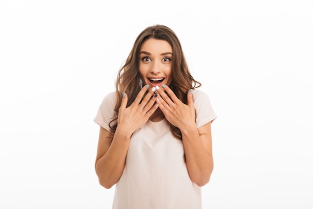 Surprised cheerful brunette woman in t-shirt covering her mouth  over white wall