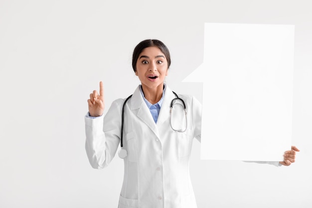 Surprised cheerful millennial indian woman doctor in coat holds abstract blank space for words and shows finger up