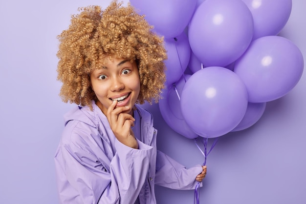 Surprised cheerful woman with curly blonde hair bites finger
looks gladfully at camera wears jacket holds bunch of inflated
balloons celebrates special occasion isolated over purple
background