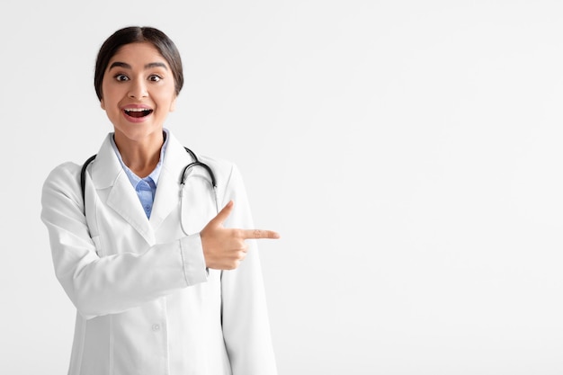 Surprised cheerful young indian woman in white coat with open mouth shows finger to empty space