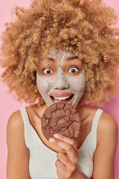 La giovane donna allegra sorpresa con i capelli ricci morde un delizioso biscotto al cioccolato si sottopone a procedure di bellezza applica una maschera nutriente vestita con una maglietta casual pone al chiuso procedure di cura della pelle