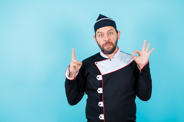 Surprised chef is looking at camera by showing a minute and okay gesture on blue background