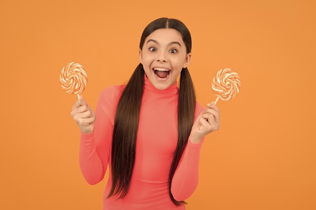 Surprised child with lollipop candy on stick on orange background childhood