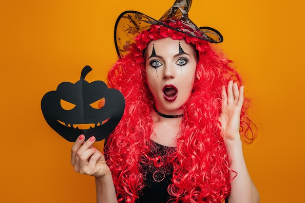 Surprised clown girl in halloween costume holds a paper pumpkin and shouts