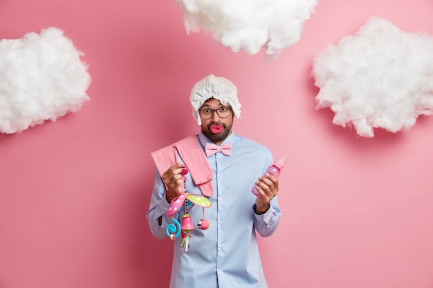 Surprised clueless bearded man prepares to become father holds feeding bottle and mobile toy sucks nipple wears diaper on head collects items for maternity hospital poses against pink wall