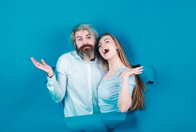 Surprised couple of bearded man and happy woman making hole in paper looking through hole advertising board blue background couple through hole in blue paper copy space for text or slogan