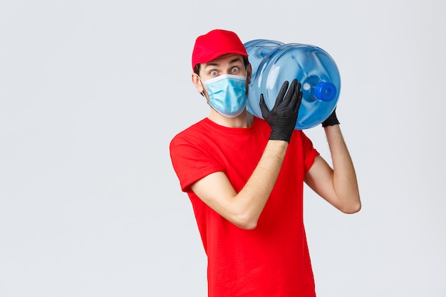 Surprised courier in red uniform cap, gloves and face mask, staring shocked as bring bottled water to office or home