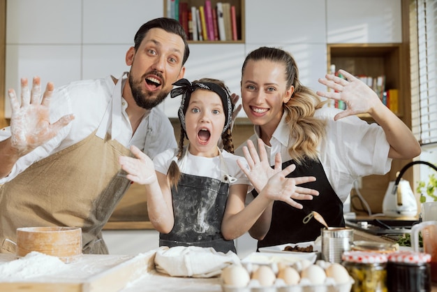 Surprised and curious daughter with young parents in aprons kneading dough for baking homemade pizza