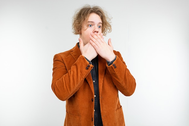 Surprised curly blond guy in a brown jacket with open mouth on a white studio background