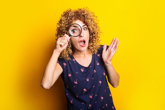 Surprised curly girl looks at the camera through a magnifying glass on a yellow surface