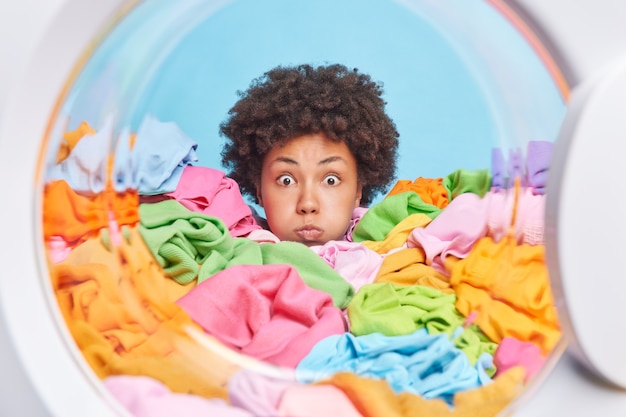 Surprised curly haired Afro American woman blows cheeks makes funny grimace drowned in multicolored laundry poses from inside of washing machine cannot believe her eyes